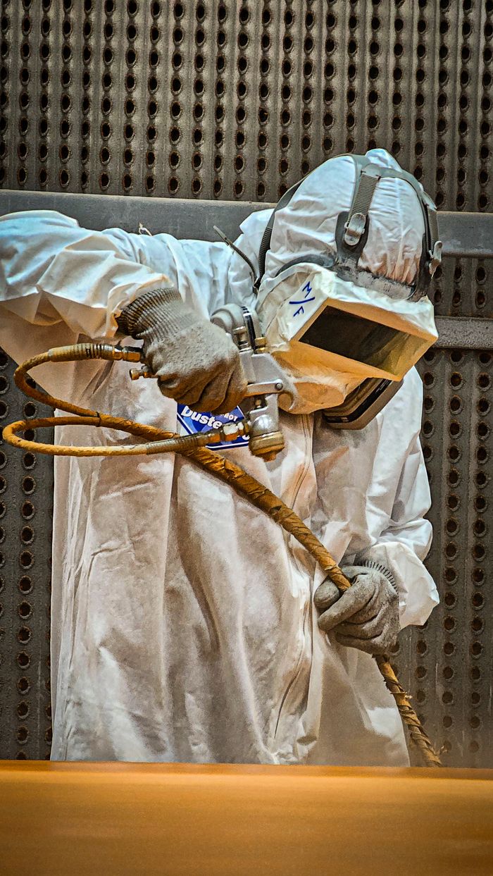 An industrial worker wearing full protective gear operates sandblasting equipment for surface treatment.